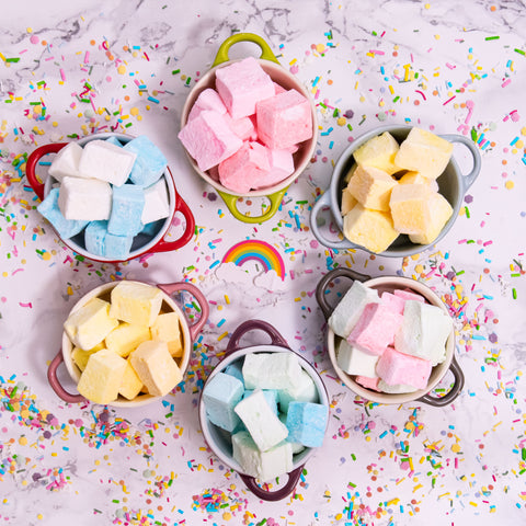 Different flavoured and coloured marshmallows sat in decorative cups on a surface with sprinkles all around
